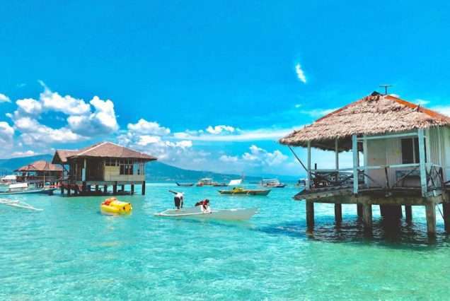 Overwater huts and boats in crystal-clear tropical sea under blue sky - highlight of Bais Dolphine Watching & Majuyod Sandbar.